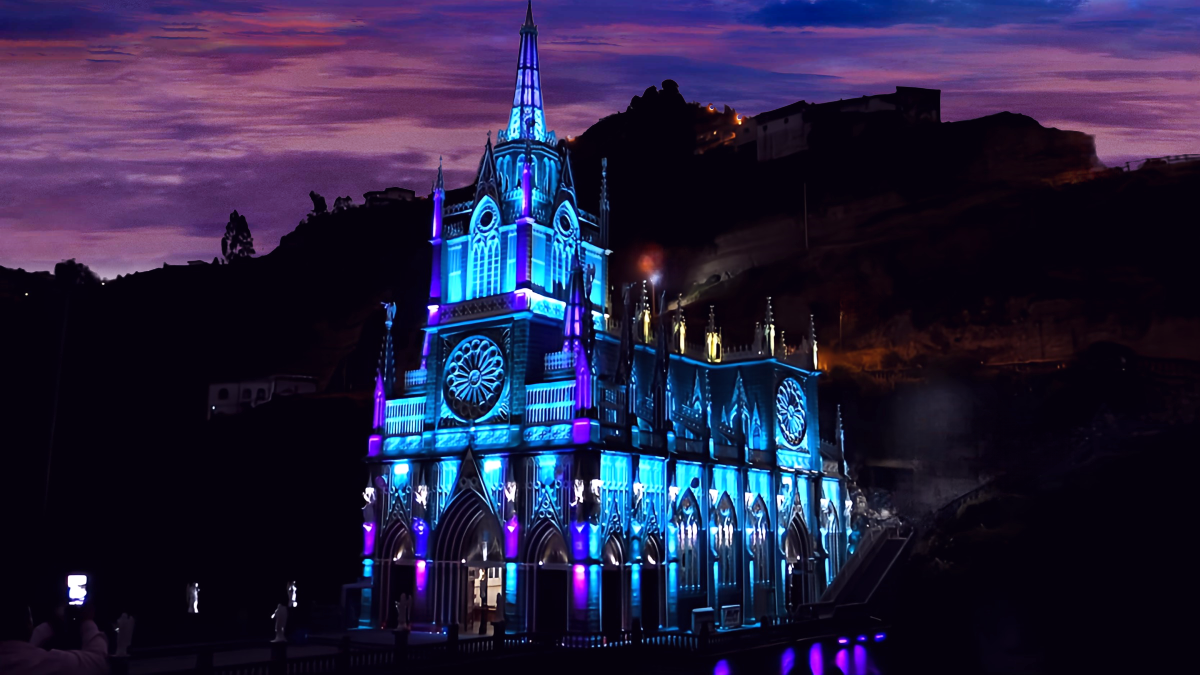 A nighttime view of Las Lajas Sanctuary illuminated in blue. The Gothic architecture glows ethereally against the dark canyon, creating a dreamlike atmosphere.